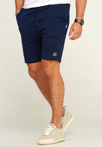 Marineblaue Shorts mit elastischem Bund und seitlichen Taschen. Mit einem weißen Logo auf dem linken Oberschenkel. Getragen mit hellen Sneakers.