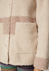 Flauschige crèmefarbene Jacke mit rosa Akzenten. Verfügt über eine strukturierte Oberfläche, einen Knopfverschluss und eine große Fronttasche.