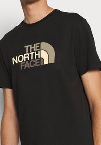 Black cotton T-shirt featuring "THE NORTH FACE" logo in beige and brown, with a rounded design element above the text.