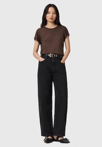 T-shirt marrone con texture, jeans neri a vita alta e gamba larga, cintura nera con fibbia dorata e scarpe basse nere. Sfondo semplice.