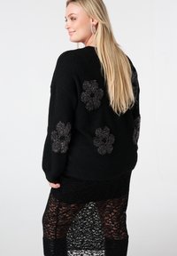 Pull noir avec des applications florales texturées sur les épaules, associé à une jupe en dentelle noire transparente avec une fente haute.