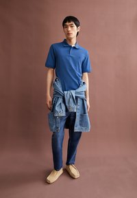 Blaues Poloshirt, Jeansjacke um die Taille gebunden, dunkle Jeans und beigefarbene Schuhe. Schlichtes Design mit kurzen Ärmeln und Kragen.