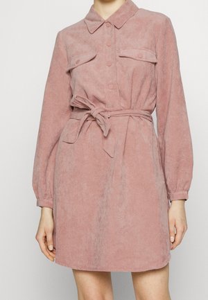Woman wearing a dusty pink corduroy shirt dress with long sleeves, buttoned front, chest pockets, and tied waist belt.