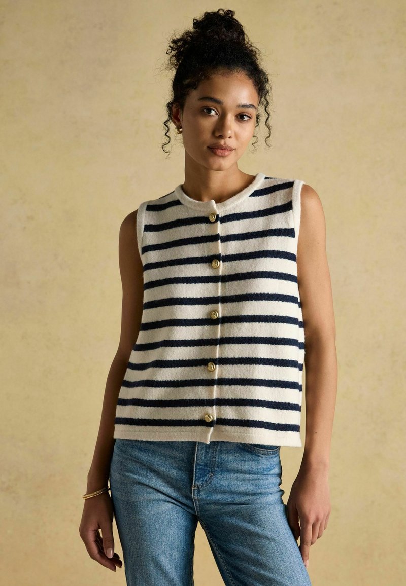 Marineblauwe en witte gestreepte mouwloze gebreide vest met gouden knopen, gedragen over lichtblauwe jeans. Zachte textuur en iets cropped ontwerp.