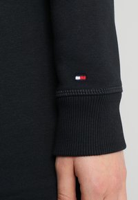 Czarny longsleeve z wywijanym mankietem o prążkowanej teksturze, z małym haftowanym logo w kolorze czerwonym, białym i niebieskim. Miękka tkanina.