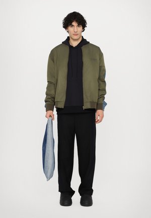 Olivgrön bomberjacka med en svart hoodie, svarta byxor och en denimväska. Slät textur med en avslappnad passform och dragkedjedetaljer.