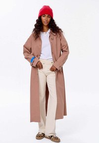 Kobieta w długim beżowym płaszczu, białej koszuli, kremowych spodniach, czerwonej czapce i butach w panterkę, stoi z rękami blisko kieszeni.