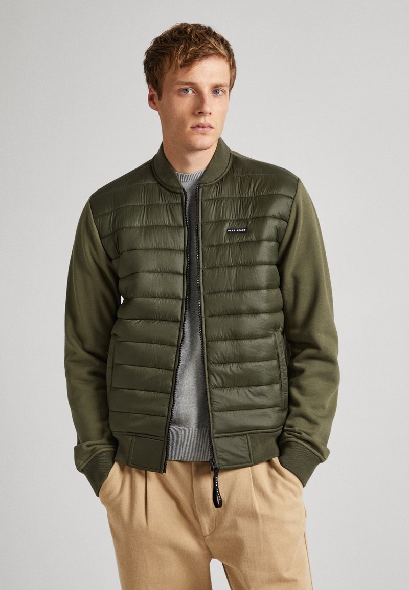 Veste bomber vert olive avec texture matelassée sur la partie supérieure, poignets côtelés, et fermeture éclair. Portée sur un pull gris.