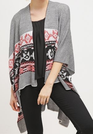 Kobieta ubrana w szary wzorzysty ponczo z różowo-czarnymi geometrycznymi motywami, na czarnej bluzce i czarnych spodniach.