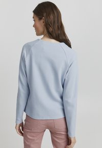 Svetlo modra majica z dolgimi rokavi, teksturirana tkanina, okrogla gola in raglan rokavi; v kombinaciji z roza hlačami. Gladek in sproščen kroj.