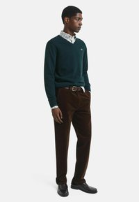 Dunkelgrüner V-Ausschnitt-Pullover über einem karierten weißen und blauen Hemd, kombiniert mit braunen Cordhosen und schwarzen Lederschuhen.