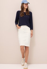 Jupe en denim blanche, mi-longue, avec des poches. Associée avec un pull bleu marine et une casquette bleu clair. Des baskets beiges avec des accents bleus complètent la tenue.
