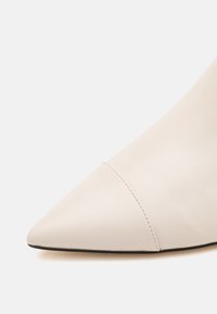 Chaussure blanche à bout pointu avec une couture près de la pointe et une semelle plate noire visible sur le bord inférieur.