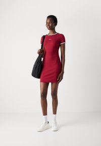 Red fitted dress with short sleeves, black and white trim, and a small logo on the chest. Paired with white sneakers and a black bag.