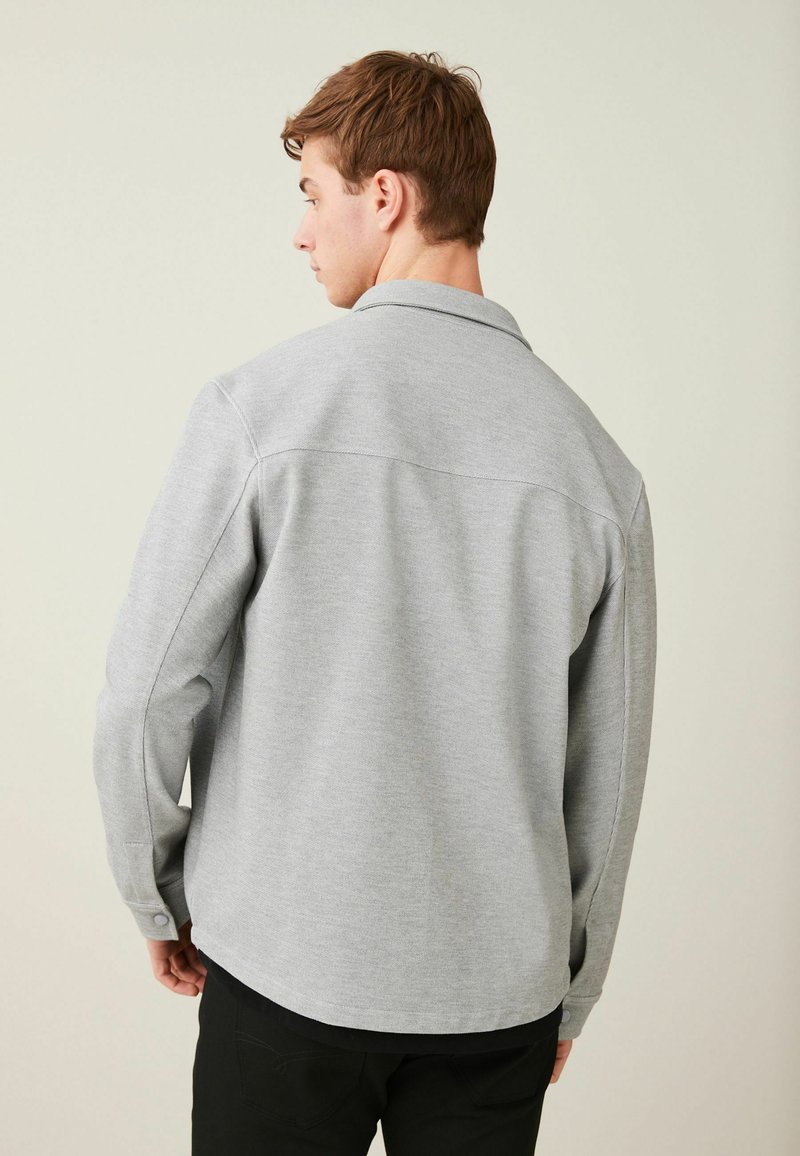 Camisa gris de manga larga con cuello; cuenta con puños abotonados y un dobladillo recto. Tela suave con una textura sutil.