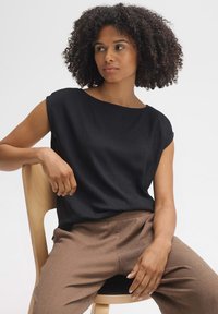 Schwarzes kurzärmeliges Oberteil aus strukturiertem Stoff mit weitem Ausschnitt. Kombiniert mit braunen, strukturierten Hosen. Modell sitzt auf einem Holzstuhl.