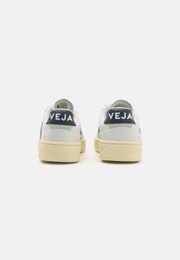 Sneakers in pelle bianca con accenti grigi, caratterizzati da una suola texturizzata, forma tondeggiante e prominente marchio "VEJA" sul tallone.