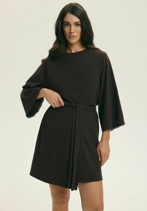 Kobieta w czarnej sukience kopertowej do kolan z szerokimi rękawami i koronkowym wykończeniem, wiążąca pasek w talii, na jednolitym tle.