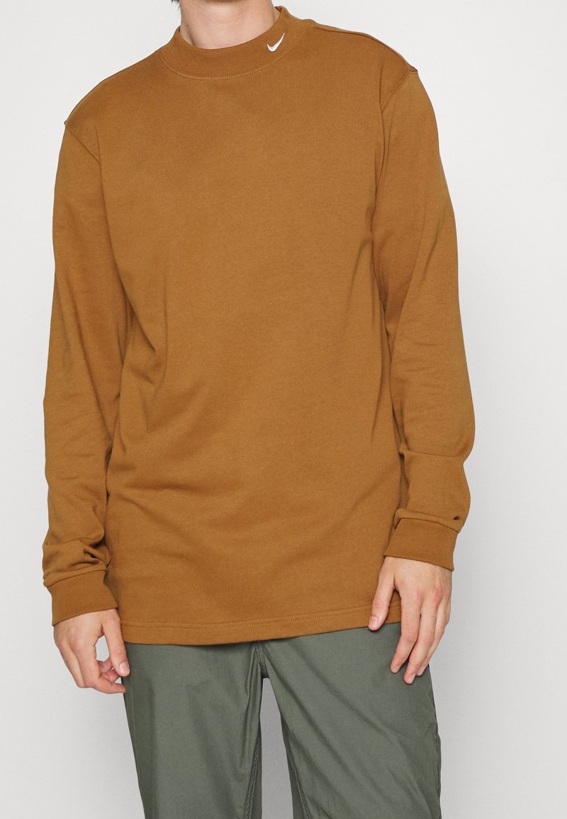 Muž oblečený v hnědém mikině s dlouhým rukávem značky Nike a šedých kalhotách, stojící proti jednoduchému světlému pozadí.