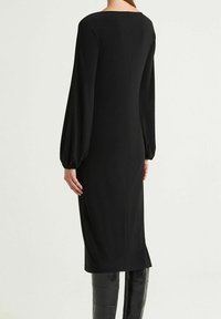 Abito nero con maniche lunghe a sbuffo e silhouette dritta. Tessuto liscio, lunghezza fino a metà polpaccio e un dettaglio sobrio di cucitura sulla schiena.