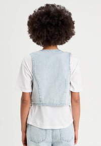 Gilet en denim avec des rayures verticales bleues et blanches, bords effilochés et design court, porté par-dessus un t-shirt blanc uni.