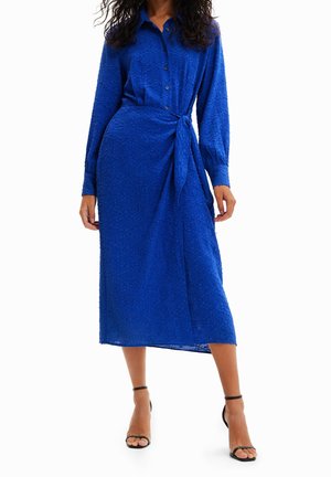 Kvinde iført en struktureret koboltblå midi skjortekjole med lange ærmer, knapper foran, bindebånd i siden og sorte ankelremssandaler.