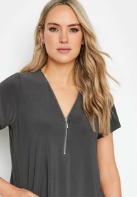 Top grigio a maniche corte con scollatura a V e dettaglio con zip sul davanti, realizzato in tessuto morbido con una vestibilità rilassata. Testura dei capelli naturale visibile.