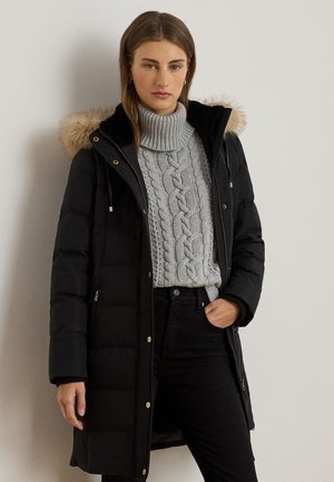 Naine, kes kannab musta pika triibulise tepitud mantlit karvaga kapuutsiga ja halli punutud kõrge kaelusega kampsunit, seisab ühe käega taskus.