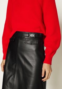 Femme portant un pull en tricot rouge rentré dans une jupe en cuir noire avec ceinture à boucle métallique, vue des épaules jusqu'à mi-cuisse.
