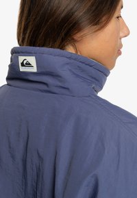 Navy blue lightweight jacket with a high collar, featuring a logo patch at the back. Smooth texture and simple design.