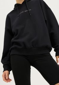 Sweat à capuche noir avec une poche kangourou à l'avant, des cordons de serrage et un texte blanc "CELESTIAL RHAPSODY". Tissu doux et poignets côtelés. Porté avec un legging noir.