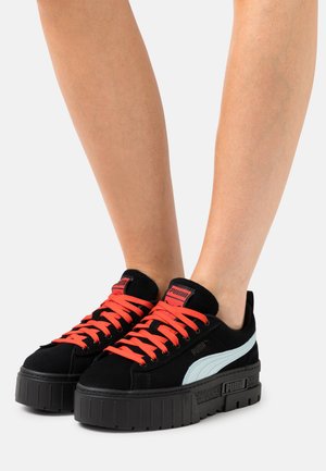 Sorte ruskinds sneakers med livlige orange snørebånd, der har en tyk sort platformssål og et lyseblåt accent på siden. PUMA-logoet er synligt.