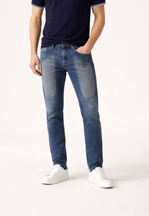 Uomo che indossa jeans blu con chiazze chiare sulle cosce, sneaker bianche e una maglietta a maniche corte blu navy, in piedi con una mano in tasca.