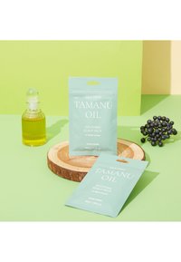 Flaska med tamanuolja i klart glas med en ribbad design, omgiven av två mintgröna påsar märkta "Soothing Scalp Pack" på en träskiva.