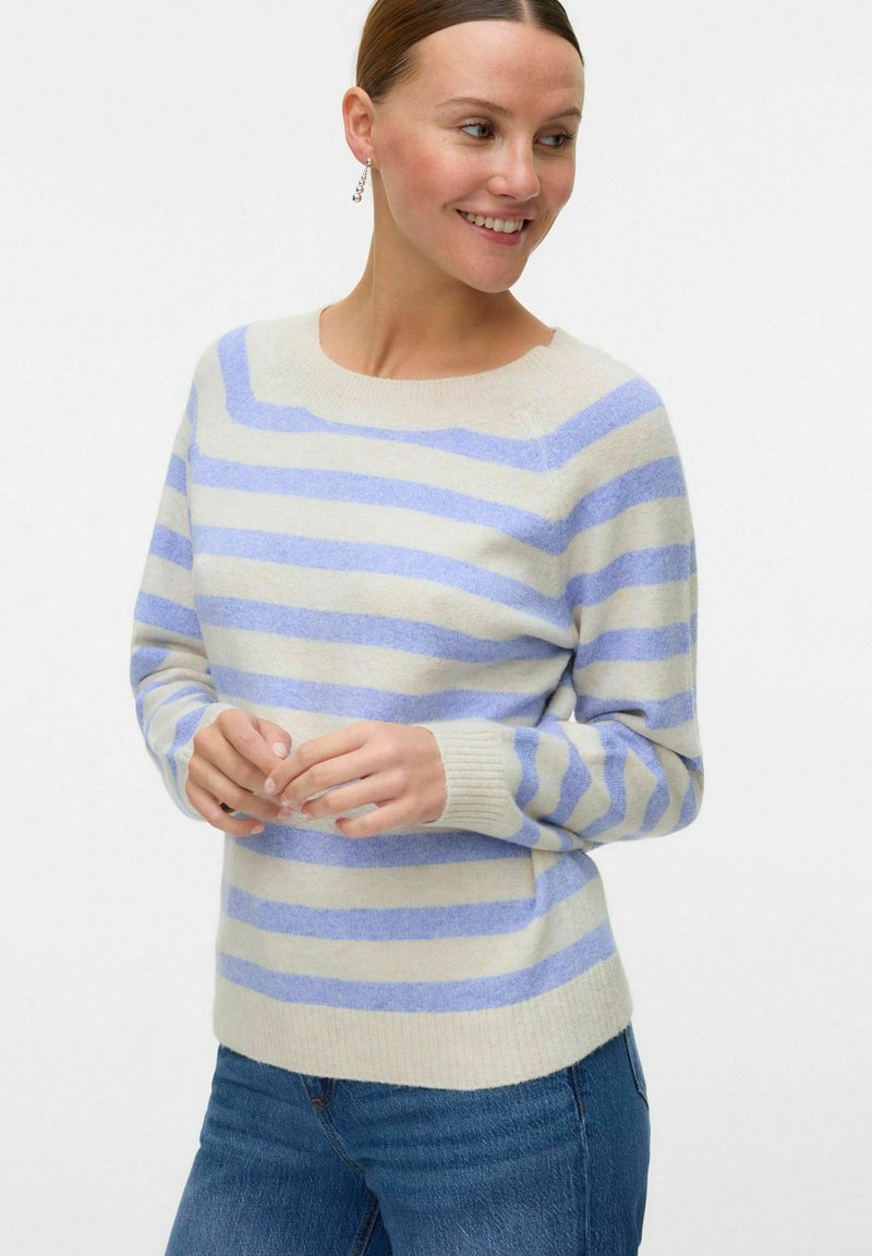 Maglione beige chiaro con righe orizzontali blu, collo a giro e polsini a coste. Texture morbida abbinata a una vestibilità casual. Jeans di denim visibili.