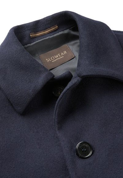 Collare di un cappotto in lana blu navy con bottone nero, che mostra l'etichetta interna con la scritta "SLOWEAR MONTEDORO" su una fodera grigia.