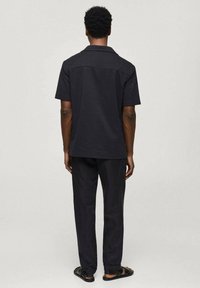 Uomo in piedi di spalle, indossando una camicia nera a maniche corte, pantaloni neri e sandali neri su uno sfondo grigio chiaro uniforme.