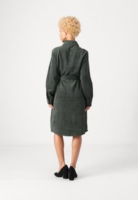 Πράσινο φόρεμα από κοτλέ με ζώνη στη μέση, μακριά μανίκια και κατακόρυφες ραβδώσεις. Μοντέλο που φοράει μαύρες ψηλές γόβες, σε θέα από πίσω.