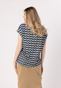 Blusa a maniche corte con un motivo ondulato nei colori blu navy, giallo e bianco, realizzata in tessuto leggero. Abbinata a una gonna beige.