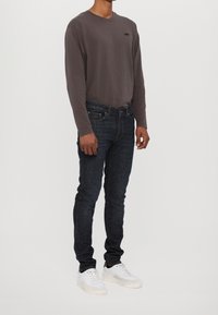 Szara bawełniana koszula z długim rękawem z małym logo, zestawiona z ciemnymi dżinsami i białymi trampkami, prezentowana w pionie na jednolitym tle.