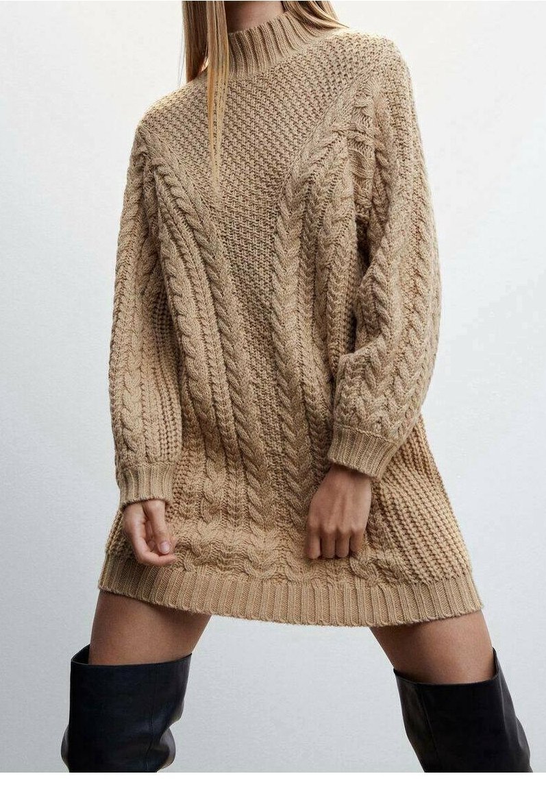 Femme portant une robe-pull beige en maille épaisse avec des motifs torsadés, associée à des bottes noires montantes, sur un fond clair uni.
