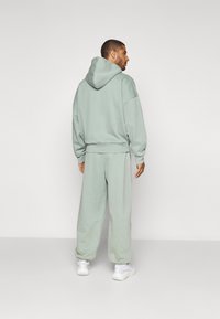 Ljusgrön hoodie med framficka, lös passform, matchas med byxor i samma färg. Tyget ser mjukt och texturerat ut. Vita sneakers syns.