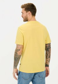 Gul kortærmet bomulds T-shirt med rund hals. Bagudvisning viser et enkelt design og tatoveringer på underarmen. Denim jeans er synlige.