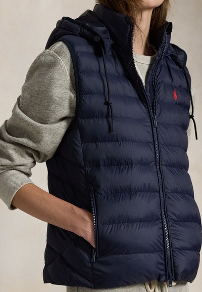 Vrouw die een marineblauw doorgestikt mouwloos vest met capuchon draagt en een grijze trui met lange mouwen, hand in de zak van het vest, tegen een effen achtergrond.