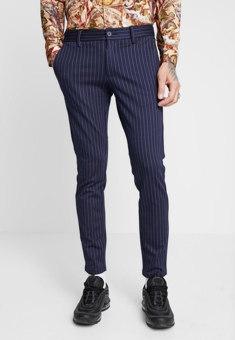 Námořnické kalhoty s proužky v elegantním, vypasovaném střihu a poutky na pásek, kombinované s černými teniskami a bílými ponožkami. Hladká textura látky.