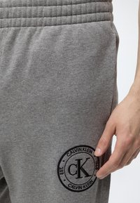 Κοντινή λήψη γκρι φόρμας με μαύρο λογότυπο Calvin Klein κοντά στην τσέπη και το δεξί χέρι ενός ατόμου που ακουμπά δίπλα.