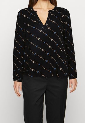 Frau trägt eine schwarze Langarmbluse mit blauen und beigen diagonalen Blumenstreifen und schwarze Hose, steht vor einfachem Hintergrund.