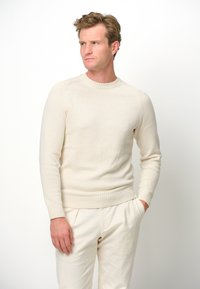 Hellbeige Strickpullover mit Rundhalsausschnitt, gerippten Bündchen und Saum sowie glatter Textur; getragen mit hellbeigen Hosen vor einem schlichten Hintergrund.