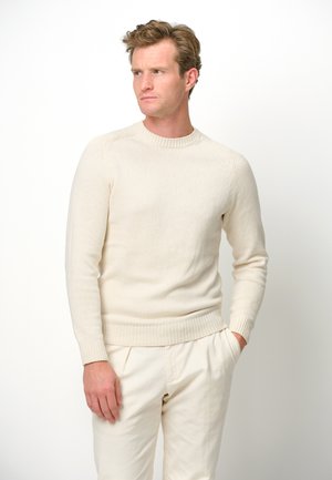 Maglione a maglia beige chiaro con scollo a giro, polsini e fondo a costine, e texture liscia; indossato con pantaloni beige chiaro su uno sfondo neutro.