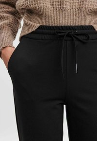 Pantalon de jogging noir avec taille élastique, fermeture par cordon de serrage et poches latérales. Le tissu semble lisse avec une coupe ajustée et un minimum de détails.
